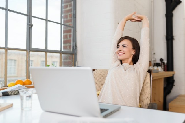 Happy casual young woman sitting in her kitchen with a laptop in front of her stretching her arms over her head and looking out the window with a smile