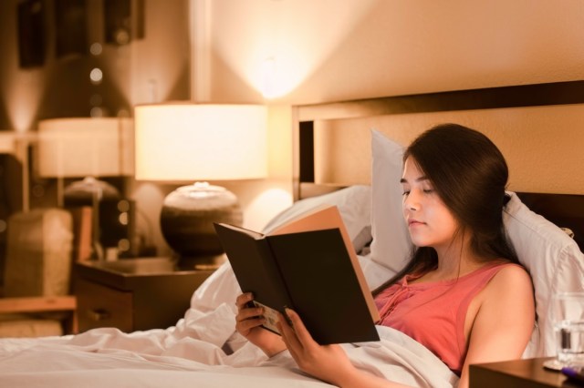 asian caucasian teen girl reading book in bed at night with yellow lamp light on walls