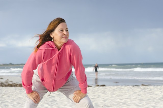 portrait of confident, healthy and sporty fit attractive looking mature woman in pink sweater, at beach, with isolated storm clouds and wild ocean as background and copy space.