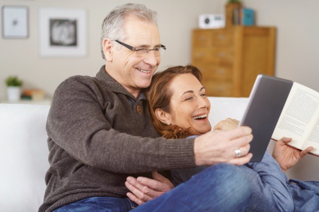 affectionate middle-aged couple relaxing on a sofa together at home laughing at something on a tablet computer, natural and spontaneous