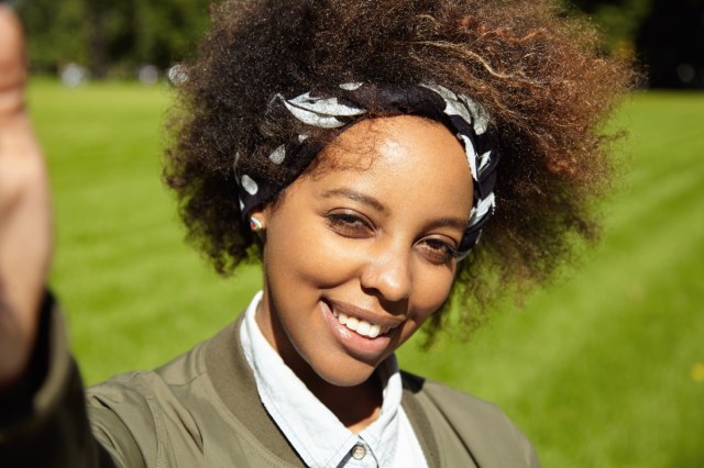 woman with afro haircut wearing black bandana, taking selfie, holding mobile phone or other device in right hand, smiling and squinting eyes in bright sun