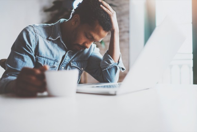 tired young african man using laptop while sitting at the table on a sunny morning. concept of people working hard at home.