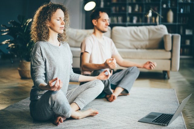 calm couple in pajamas meditating, listening spiritual practice lessons on laptop, sitting on lotus stand at home