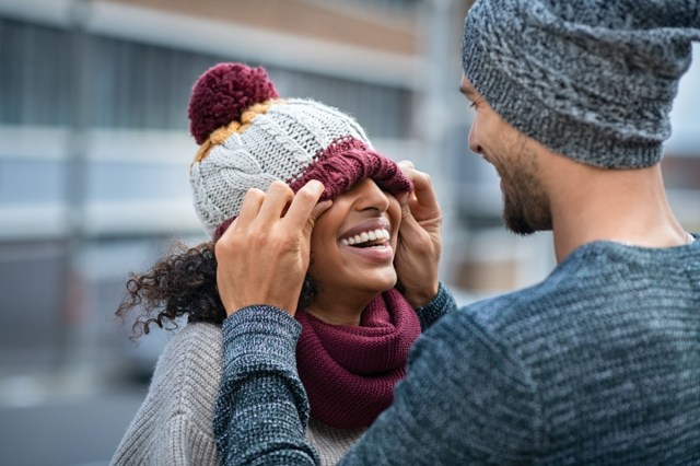 couple having fun outdoors in the winter.  young man covering eyes his girl with wool cap