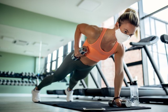 female athlete with protective face mask doing plank exercise with hand weights in a gym.