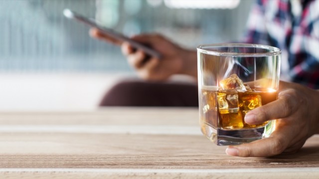 man relaxing with bourbon whiskey drink alcoholic beverage in hand and using mobile smartphone