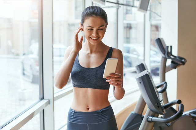 woman in sportswear choosing music for fitness on her smartphone while standing against treadmills at the gym