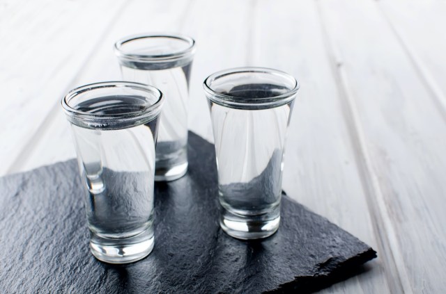 three vodka shots with ice on black stone board on white wooden table, copy space