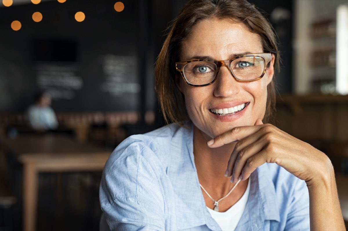 portrait of happy mature woman wearing eyeglasses and looking at camera.  close-up face of smiling woman sitting in cafeteria with hand on chin.  successful lady in a cafe pub.