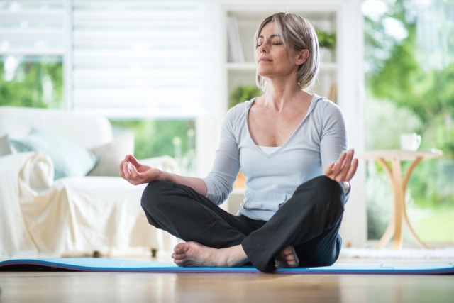 middle aged woman sitting in lotus position on a rug in his living room.  her eyes are closed.  she is in the foreground