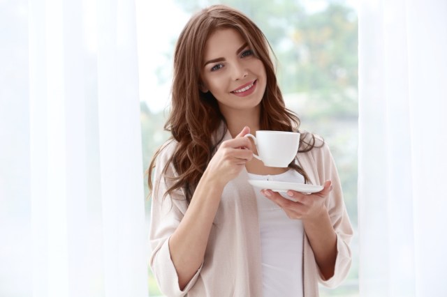 woman holding a cup of coffee standing near window.