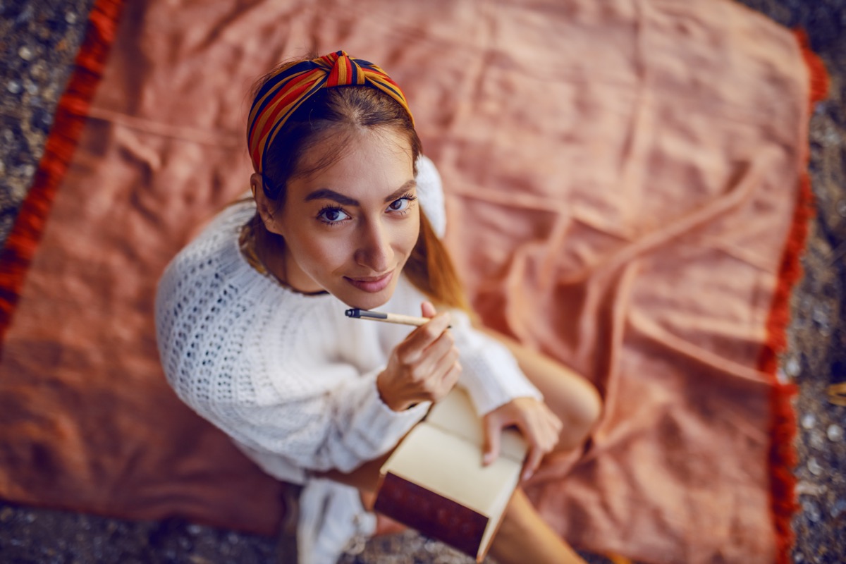 top view of caucasian brunette in sweater and with headband sitting on blanket outdoors and holding pen and diary in hands while looking at camera.