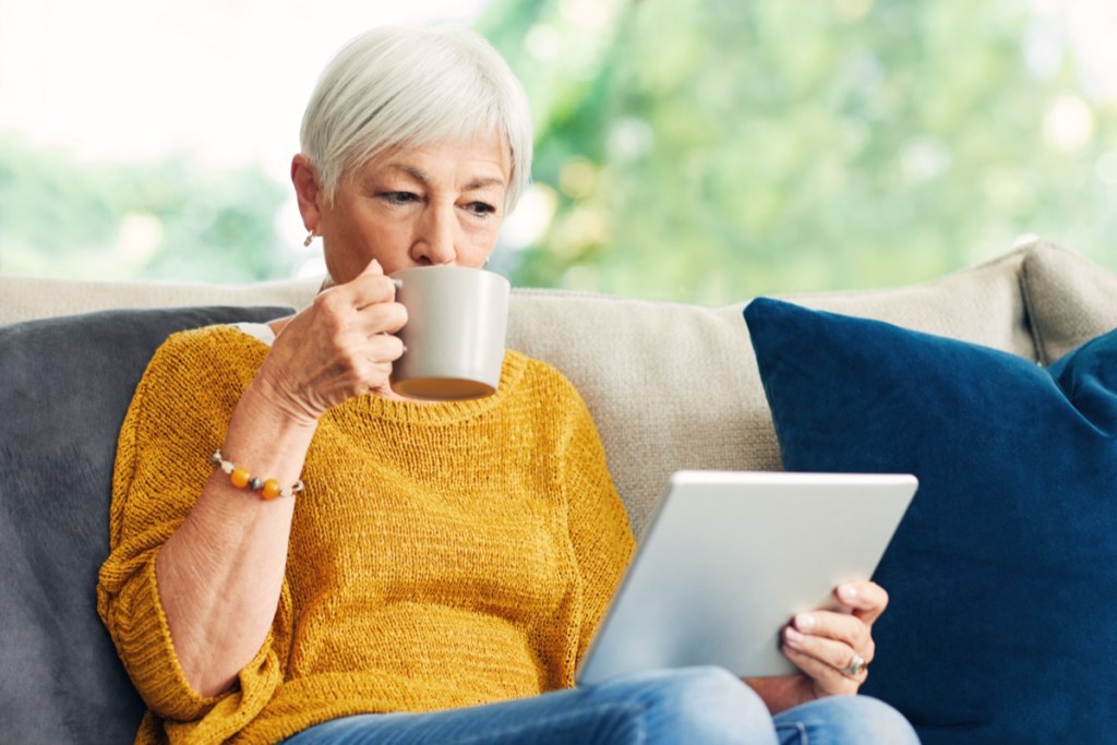 senior woman using a digital tablet at home and having coffee on the couch.