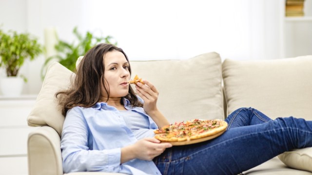 woman is chewing pizza, while laying on the white sofa. she is watching tv shows, being on blurred background.