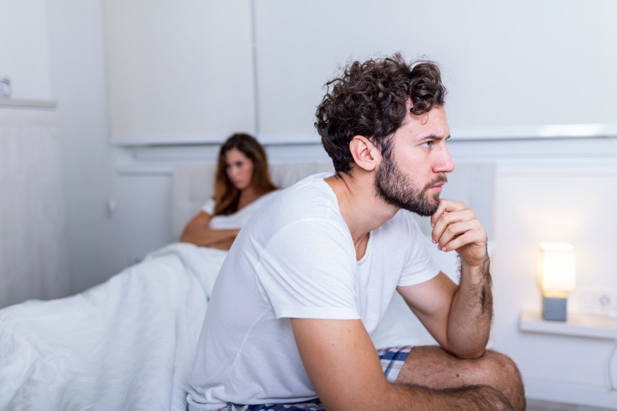 sad man sitting on a bed, girlfriend in the background.