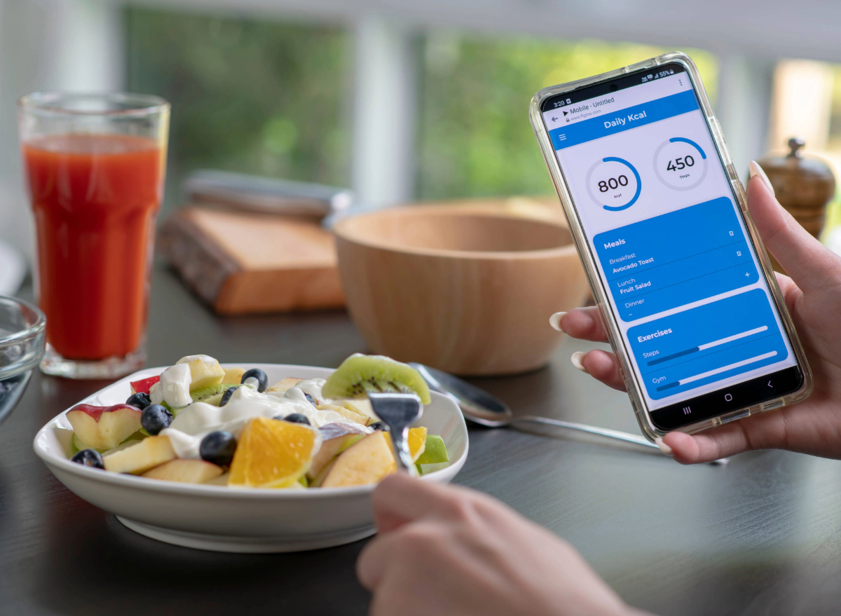 close-up of woman holding phone in hand while eating fruit salad and tracking her calorie count