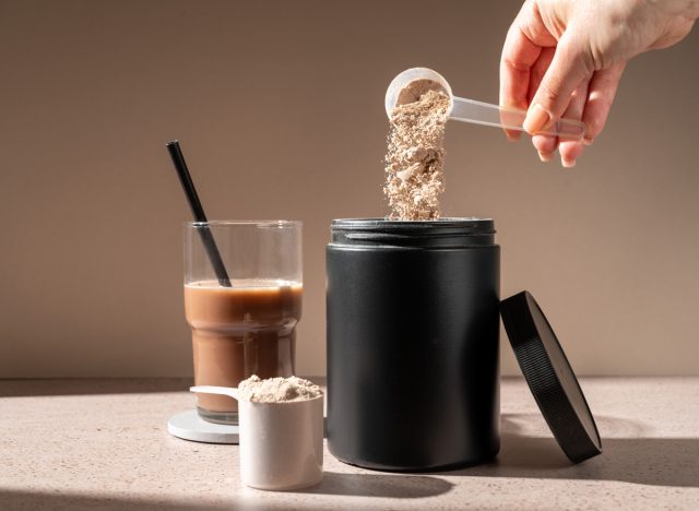 woman holding a scoop of protein powder above the container with a protein shake in the background