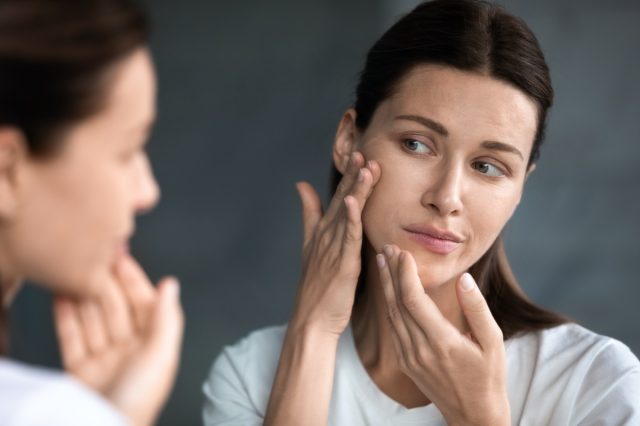 woman looking at red acne spots on chin in mirror, upset young woman unhappy with unhealthy skin