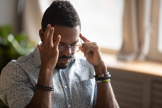 african ethnicity guy sitting indoors eyes closed touching temples with fingers suffering from migraine feeling unhealthy. student is preparing for exams or trying to remember forgotten important information