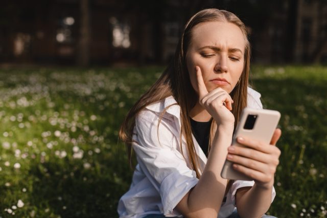 woman holding a phone while resting her chin on her hand. expressive student thinking outside on campus.