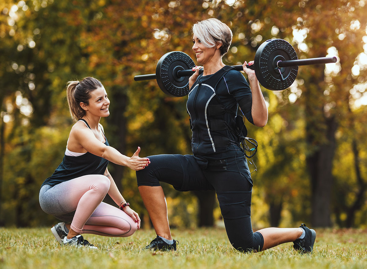 a mature woman does lunges exercises with a personal trainer in the park, wearing a black suit with an ems electronic simulator to stimulate the muscles.